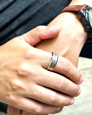 Anel masculino de aço bicolor nas cores prata e rosé da UNA Semijoias no dedo, em close das mãos com relógio de couro marrom e look casual.