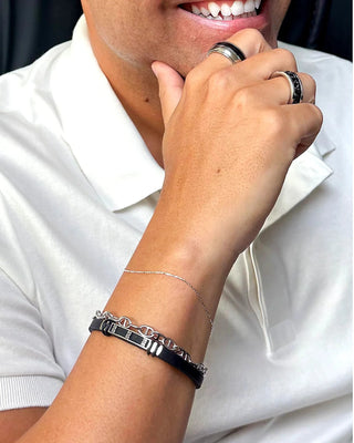 Anel masculino de aço inox 316L com faixa preta central da UNA Semijoias no dedo, em close do sorriso e do braço com pulseira masculina sobre camisa branca.