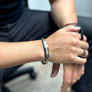 Bracelete de aço bicolor masculino rígido com placa da UNA no pulso, acessório sofisticado em foto editorial