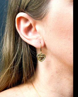 Brinco coração Espírito Santo folheado a ouro 18k da UNA Semijoias na orelha feminina em close lateral, com detalhes em relevo dourado.