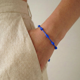 Pulseira verão nozinho da sorte azul da UNA Semijoias no pulso feminino, em close lateral, combinada com look casual em tecido claro.